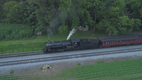 Vista-Aérea-De-Un-Motor-De-Vapor-De-1924-Con-Un-Tren-De-Pasajeros-Que-Viaja-A-Lo-Largo-De-La-Campiña-Amish-Mientras-Las-Puestas-De-Sol-En-Un-Día-De-Verano-Visto-Por-Un-Dron