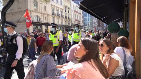 People-eating-outside-a-restaurant-watch-protesters-marching-against-lockdowns-and-vaccine-passports-along-Oxford-Street