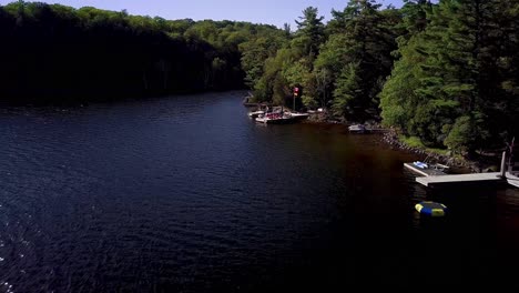 Drone-Flying-over-Cottage-Docks-on-Lake,-Canada-and-Germany-Flags-Flying-in-Wind-and-Green-Pine-Trees-on-Shore