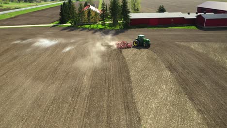 Folgende-Luftaufnahme-Eines-Grünen-Doppelradtraktors-Mit-Landwirtschaftlichen-Gebäuden-Im-Hintergrund,-Der-Im-Frühling-Ein-Landwirtschaftliches-Feld-Mit-Einem-Roten-Grubber-Bearbeitet