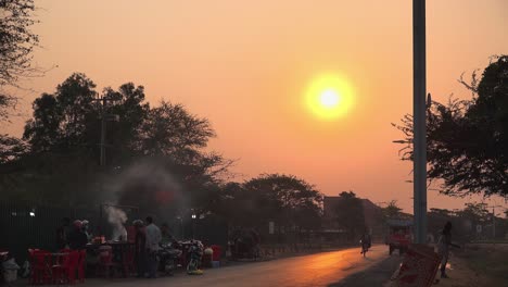 Lapso-De-Tiempo-De-Una-Puesta-De-Sol-En-Una-Calle-Concurrida-Con-Un-Puesto-De-Comida-Callejera-Preparando-Comida-Y-Vendiéndola-En-La-Carretera-Y-Muchos-Vehículos-Pasando