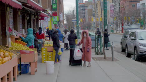 Ein-Maskierter-Fußgänger-Zieht-Einen-Koffer-Die-Straße-Entlang,-Vorbei-An-Einem-Freiluftmarkt-In-Chinatown