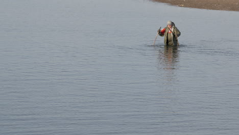 Old-fisherman-with-wooden-sticks-walking-through-water-of-ocean-after-exhausted-fishing-day