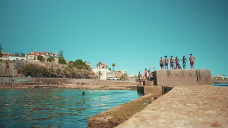 Salt-water-swimming-pool-at-shoreline-with-kids-jumping-from-pier-on-water-at-sunshine-wide-angle-4K