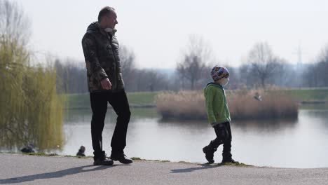 Niño-Con-Mascarilla-Caminando-En-El-Parque-Junto-Al-Lago-Con-El-Padre-En-La-Mañana-Soleada