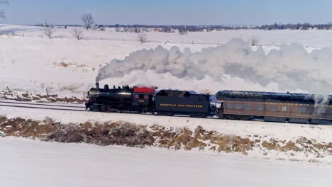 Vista-Aérea-De-Una-Antigua-Locomotora-De-Vapor-Que-Se-Acerca-Tirando-De-Automóviles-De-Pasajeros-Y-Soplando-Humo-Y-Vapor-Después-De-Una-Tormenta-De-Nieve