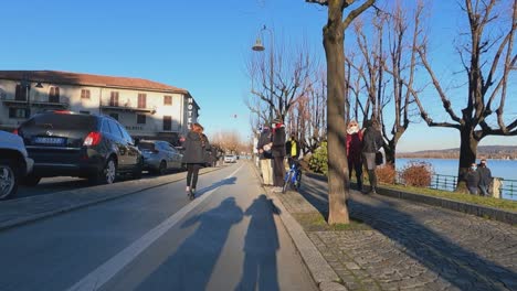 Ego-Tracking-Ansicht-Eines-Kleinen-Mädchens,-Das-Spaß-Beim-Fahren-Eines-Elektrorollers-Auf-Dem-Fahrradweg-Am-Wasser-Von-Arona-Hat