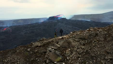 Touristen,-Die-Auf-Einem-Berg-Gegen-Fagradalsfjall-Mit-Lava-Und-Rauch-Unter-Einem-Bewölkten-Himmel-In-Island-Spazieren---Drohnenaufnahme-Aus-Der-Luft