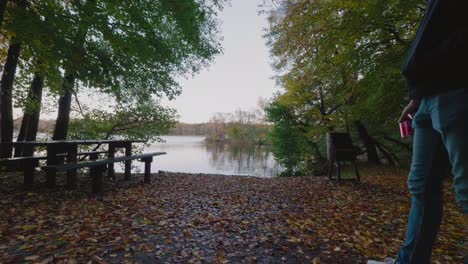 Un-Tipo-Alto-Con-Gorra-Sosteniendo-Latas-De-Soda-Camina-Y-Mira-El-Lago-Gyllebo-En-Otoño,-Österlen-Suecia---Plano-General-Estático