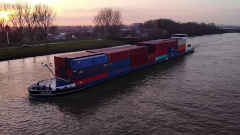 Luftaufnahme-Gestapelter-Container-Auf-Dem-Frachtschiff-Julia-Entlang-Der-Oude-Maas-Vor-Gelbem-Sonnenuntergangshimmel