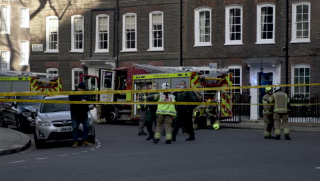 Un-Equipo-De-Bomberos-En-La-Escena-De-Un-Incendio-En-Smiths-Square-Londres,-Un-Bombero-Que-Lleva-A-Los-Investigadores-A-Un-Lugar-Seguro-En-El-Perímetro-Que-Permite-Que-Los-Servicios-De-Emergencia-Hagan-Su-Trabajo,-Inglaterra