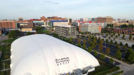 Aufsteigende-Luftaufnahme-Des-Lexus-Velodroms-Während-Der-Goldenen-Stunde-Mit-Einer-Skyline-Der-Stadt-An-Einem-Schönen-Tag