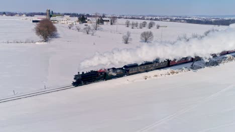 Vista-Aérea-De-Una-Antigua-Locomotora-De-Vapor-Que-Se-Acerca-Tirando-De-Automóviles-De-Pasajeros-Y-Soplando-Humo-Y-Vapor-Después-De-Una-Tormenta-De-Nieve