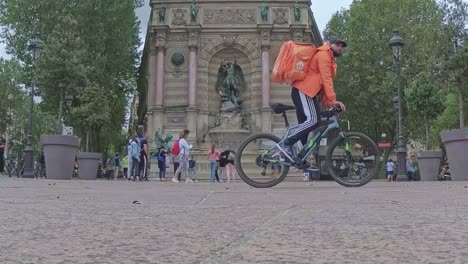 Una-Toma-En-Cámara-Lenta-De-La-Fontaine-Saint-michel-En-Las-Calles-De-París,-Con-Un-Ciclista-Naranja-Y-Gente-Caminando