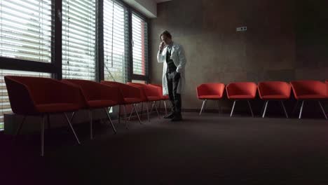 Close-up-side-view-of-a-middle-aged-Caucasian-male-doctor-standing-near-the-chair-at-an-empty-seminar-and-talking-on-a-smartphone