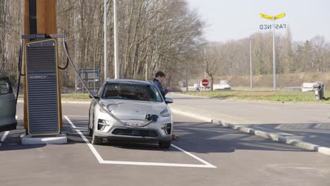 Hübscher-Geschäftsmann-Nutzt-Smartphone-Und-Stützt-Sich-Auf-Ein-Luxuriöses-Fahrzeug,-Während-Er-Darauf-Wartet,-Dass-Das-Elektroauto-Neben-Der-Autobahn-Aufgeladen-Wird---Leuven,-Belgien