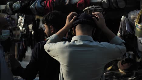 Vista-Posterior-De-Un-Hombre-Probándose-Una-Gorra-En-El-Mercado-De-Karachi,-Pakistán