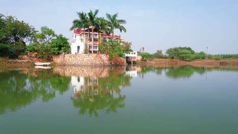 Aerial-drone-shot-forward-flying-over-the-blue-green-lake-towards-the-beautiful-white-vacation-house-with-three-floors-and-red-brick-roof-in-Vadodara,-India