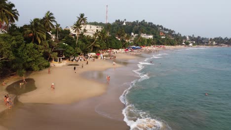 Dronie-Shot-Of-Two-Pretty-excited-Girls-Wearing-Swimsuit-Running-On-Beach-Towards-The-Sea,-Sri-Lanka