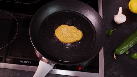 High-angle-dolly-in-shot-of-golden-brown-schnitzel-being-cooked-in-a-black-pan-and-stove-top,-with-fresh-vegetables-on-the-counter,-slow-motion