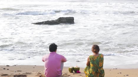 Dos-Personas-Se-Sientan-En-La-Playa-De-Arena,-Miran-El-Mar,-Las-Olas-Se-Curvan-Y-Se-Rompen