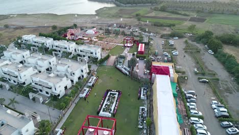 traveling-shot-covering-several-white-buildings-surrounded-by-an-event-set-up-with-colorful-stages-and-decorations,-multiple-organized-chair-set-up,-flashing-screens,-bright-outdoors,-water-visible