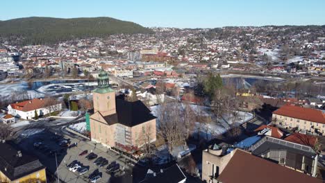 Aufsteigende-Luftaufnahme-Der-Kongsberg-Kirche---Kirche-Mitten-Im-Stadtzentrum-Mit-Dem-Fluss-Numedalslagen-Dahinter---Blauer-Himmel-Im-Winter-Norwegen