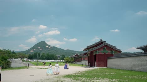 La-Gente-Entra-Por-La-Puerta-De-Yongseongmun-Al-Palacio-Gyeongbokgung,-Las-Chicas-Vestidas-Con-Hanbok-Caminan-Hacia-La-Entrada-Con-La-Cumbre-Del-Monte-Bukhansan-Al-Fondo-En-Un-Día-Nublado