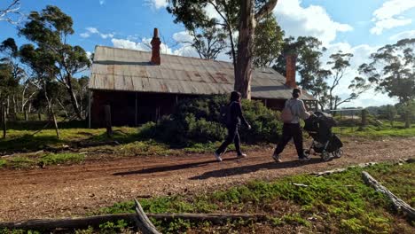 Mujer-Caminando-Por-Un-Camino-De-Tierra,-Empujando-Un-Cochecito-Más-Allá-De-Una-Antigua-Casa-Abandonada-Con-Techo-De-Hojalata-En-La-Victoria-Regional