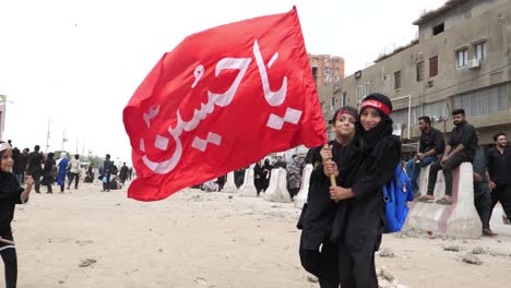 Dos-Niños,-Un-Niño-Y-Una-Niña,-Levantan-Banderas-Rojas-Durante-El-Juloos-O-Desfile-De-Muharram,-También-Conocido-Como-Muharram-Ul-Haram,-Que-Se-Considera-El-Segundo-Mes-Más-Sagrado-Después-Del-Ramadán