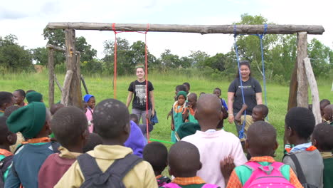 Two-Young-American-Girls-Pushing-Smiling-African-Children-on-a-Playground-Swing-at-a-Primary-School-in-Rural-Zimbabwe,-Slow-Motion-Wide-Angle