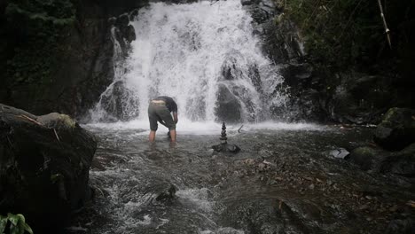 Slow-motion-tilt-up-shot-of-an-Indonesian-man-building-cairns-in-a-river-in-front-of-a-waterfall