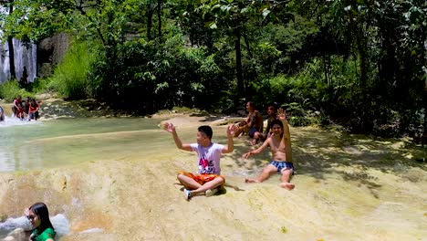 A-beautiful-waterfall-cascades-down-a-cliff-into-a-beautiful-turquoise-pool-below-in-the-middle-of-a-lush-green-jungle-in-Bohol,-Philippines-while-locals-enjoy-the-cool-relaxing-water