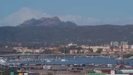 Toma-Aérea-Estable-Para-Una-Costa-De-Mar-Del-Puerto-De-La-Ciudad-Llena-De-Camiones,-Barcos,-Contenedores-De-Envío-Con-Vistas-A-Los-Edificios-De-La-Ciudad-Y-Montañas-Verdes-En-El-Fondo