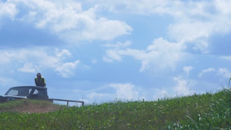 Track-safety-man-watches-how-autocross-buggies-are-jumping-over-the-hill-in-amateur-racing-on-the-dirt-track-in-sunny-summer-day,-medium-shot