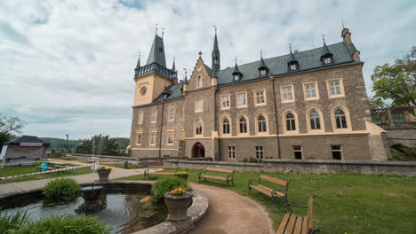 Czech-renaissance-palace-with-a-fountain-and-people-outside-with-puffy-clouds-rushing-over-turquoise-sky,-time-lapse