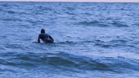 Surfer-on-surfboard-paddling-over-waves-near-the-Baltic-sea-Karosta-beach-at-Liepaja-during-a-beautiful-vibrant-sunset-at-golden-hour,-medium-shot-from-a-distance