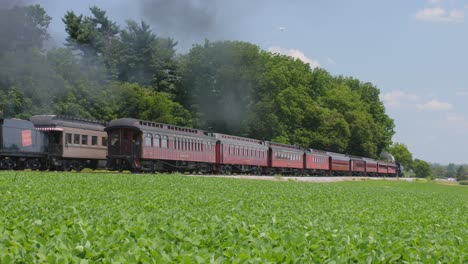 Eine-Dampflokomotive-Aus-Dem-Jahr-1910-Mit-Personenzug-Fährt-In-Einen-Bahnhof-Ein-Und-Bläst-Schwarzen-Rauch-Durch-Die-Amish-Landschaft,-Während-An-Einem-Sonnigen-Sommertag-Eine-Zweite-Dampflokomotive-Vorbeifährt