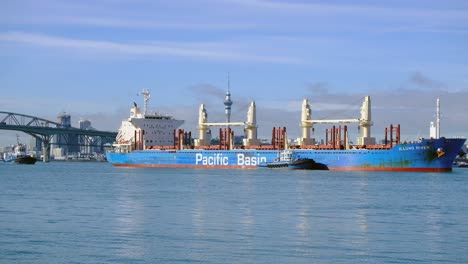 Primer-Plano-De-Un-Buque-De-Carga-Que-Viaja-A-La-Fábrica-De-Azúcar-De-Chelsea-En-El-Puerto-De-Auckland-Con-La-Ciudad-Del-Cielo-Y-El-Puente-Del-Puerto-Como-Fondo