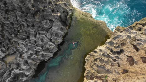 Aerial-top-view-of-man-floating-in-Angel's-billabong-natural-pool,-boom-up,-Bali