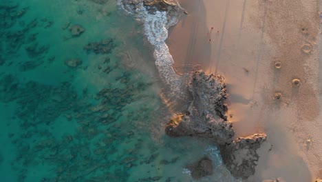 Spektakuläre-Aussicht-Von-Oben-Auf-Den-Türkisfarbenen-Paradiesischen-Ozean,-Den-Meeresboden-Und-Die-Wellen,-Die-Am-Unberührten-Sandstrand-Der-Küste-Krachen,-Mit-Menschen,-Die-Bei-Sonnenuntergang-An-Der-Küste-Entlang-Laufen,-Direkt-über-Der-Luftaufnahme