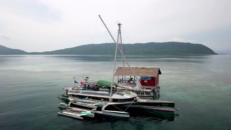 Bigkanu-trimaran-sailboat-cruise-stationed-in-Friwen-Village-with-passengers-on-deck,-Aerial-circle-around-shot