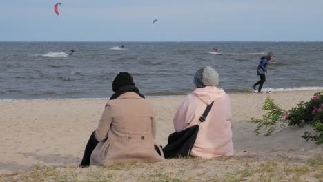 Dos-Personas-Sentadas-En-La-Arena-Mirando-A-Los-Kitesurfistas-En-El-Mar-Báltico-En-Rewa,-Polonia---Midshot