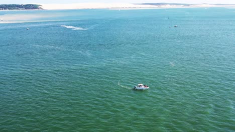 Boat-stopped-on-Arcachon-Bay-with-people-onboard-and-Dune-du-Pilat-on-the-background,-Aerial-dolly-out-reveal-shot