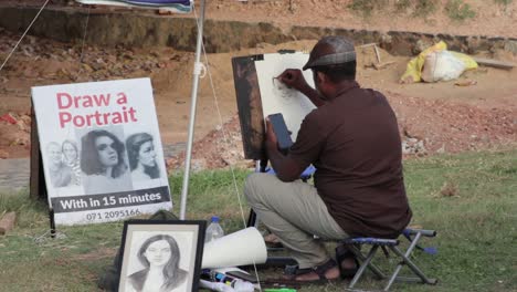 El-Artista-De-Bocetos-Callejeros-Realiza-Un-Retrato-A-Lápiz-En-Vivo-De-Un-Niño-Pequeño-En-Un-Trozo-De-Papel-Blanco,-Sentado-En-Una-Pequeña-Silla-Plegable-Bajo-Un-Gran-Paraguas,-Fuerte-Galle-Holandés,-Sri-Lanka