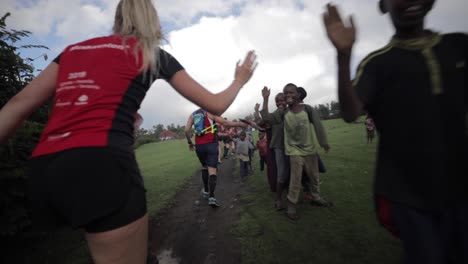 Atletas-De-Maratón-Corriendo-En-Un-Camino-De-Tierra-Seguidos-Dando-Palmas-A-Los-Niños-En-El-Camino-Y-Sonriendo-En-Los-Campos-De-La-Jungla-De-Tanzania---Cámara-Lenta