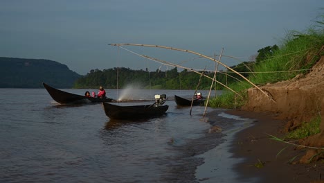 Langes-Boot-Auf-Dem-Mekong,-Mit-Einem-Piloten-In-Rot-Und-Zwei-Weiteren-Passagieren,-Der-An-Zwei-Anderen-Booten-Vorbeirast,-Die-Hüpfend-Auf-Dem-Fluss-Vertäut-Sind,-Am-Ufer-Auf-Der-Thailändischen-Seite-In-Ubon-Ratchathani