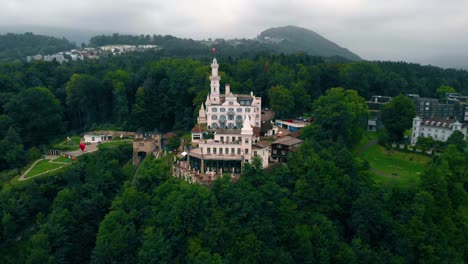 Aerial-view-away-from-a-white-castle,-restaurant-and-hotel,-on-the-Chateau-hill,-dark,-cloudy,-summer-day,-in-Lucerne,-Switzerland---pull-back,-drone-shot