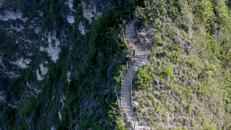 Kelingking-Beach-cliff-staircase-with-people-resting-in-the-Hinduist-shrine-area-at-the-top,-Aerial-fly-over-reveal-shot