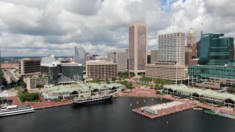 Absteigende-Luftaufnahme-Des-Innenhafens-Von-Baltimore-An-Sonnigen-Sommertagen,-Hohe-Wolkenkratzer-Und-Bauschige-Wolken-Füllen-Den-Himmel-An-Der-Touristenattraktion-Am-Wasser